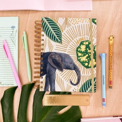Caderno com capa de elefante, folhas verdes e padrões dourados, entre material de escritório e planta verde sobre mesa de madeira