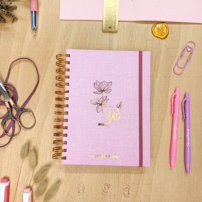 Caderno rosa 2027 com tema floral, tesoura, régua dourada e canetas coloridas numa mesa de madeira