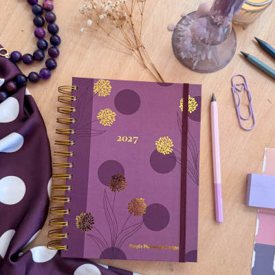 Agenda roxa 2027 com flores douradas, colar de contas, vela derretida e utensílios de escrita numa mesa de madeira.