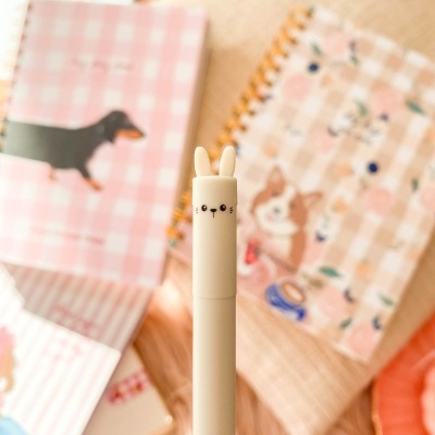 Caneta branca com tampa de coelho e texto roxo, agendas com padrões e cães ao fundo