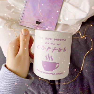 Caneca branca com design lilás e texto sobre café, segurada por uma mão sobre manta cinzenta