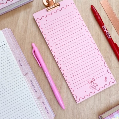 Bloco de notas rosa com canetas, régua metálica, clip e agendas numa mesa de madeira clara