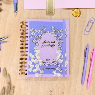 Caderno lilás espiral com design floral dourado e canetas numa mesa de madeira
