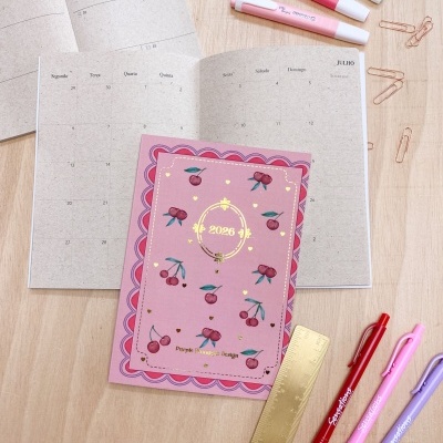 Planeador mensal 2026 com capa rosa decorada com cerejas e canetas de cores variadas numa mesa de madeira