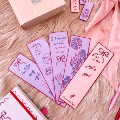 Marcadores de livros coloridos rosa e lilás com textos e desenhos, livro rosa e lenço rosa claro sobre superfície felpuda rosa.