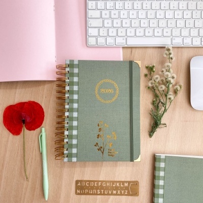 Agenda verde com espiral dourado e detalhes florais ao lado de vários objetos sobre uma mesa de madeira clara