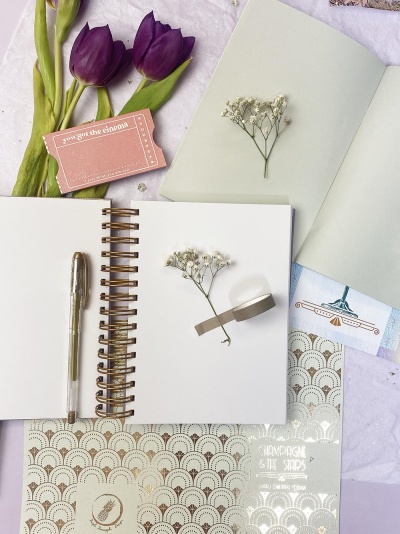 Cadernos espiralados com caneta, flores secas, cartão rosa com texto e padrão dourado sobre fundo claro