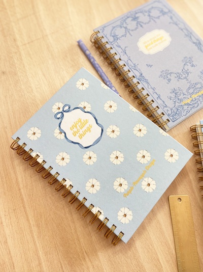 Dois cadernos azuis claros com temas florais e texto em dourado em cima de uma mesa de madeira