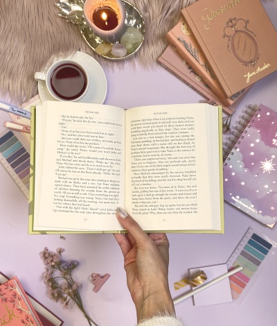 Mesa com livro aberto e vários materiais de papelaria sobre superfície lilás e pelo bege.