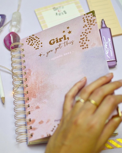 Caderno espiral rosa com texto dourado e mão feminina com anéis