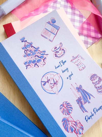 Caderno com capa decorativa com desenhos e textos em azul e rosa, tecido xadrez ao fundo
