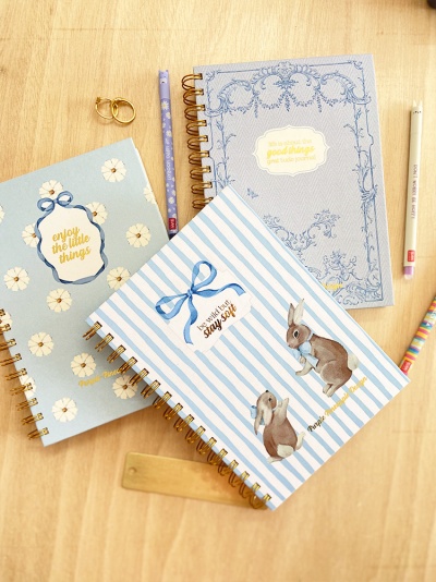 Três cadernos decorativos sobre mesa de madeira com lápis e acessórios