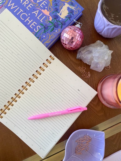 Mesa com agenda aberta, caneta rosa, livro azul, bola decorativa rosa, pedra cristal, clips rosa, vela cor-de-rosa e chá