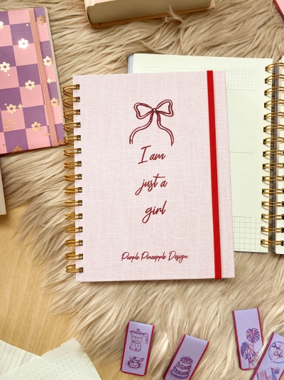 Caderno rosa claro com texto em vermelho e elástico vermelho, rodeado de outros cadernos e marcadores