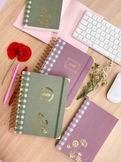 Cadernos com capas verdes e lilás com detalhes dourados e padrão xadrez, caneta rosa, flores, teclado e rato branco sobre mesa de madeira