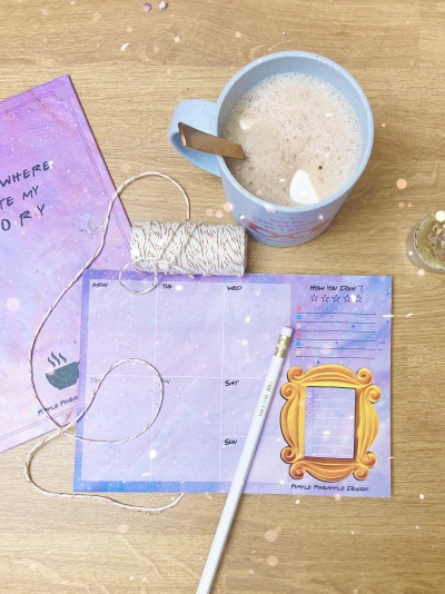 Mesa organizada com calendário semanal roxo claro, caneta branca, rolo de corda, caneca azul com bebida com colher e marcador de página roxo.