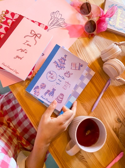 Mesa de madeira com agendas coloridas, auscultadores brancos, óculos de sol, caneta e chávena com chá.