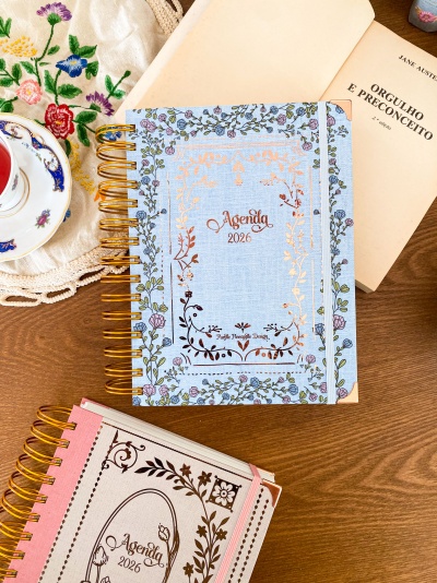 Dois cadernos com espiral dourado e capas florais, um azul claro e outro rosa claro, ambos com texto Agenda 2026.