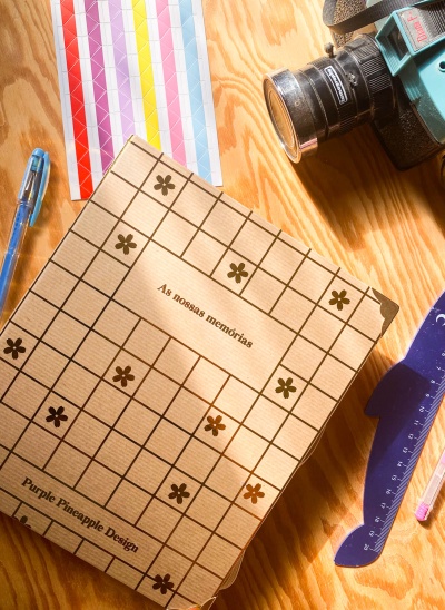 Caderno quadriculado com texto 'As nossas memórias' sobre mesa com caneta, fita métrica, régua e câmara