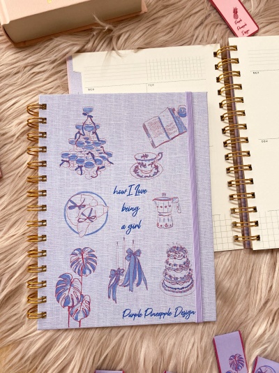 Agenda lilás com ilustrações e texto, espiral dourada e elástico, sobre superfície peluda rosa clara