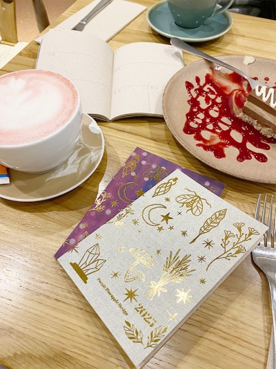 Mesa com café, fatia de bolo, duas agendas decoradas e uma caneca verde