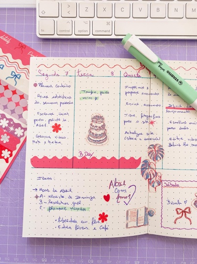 Página de planner escrita à mão com marcadores e desenhos decorativos, ao lado de marcador verde pastel e teclado branco
