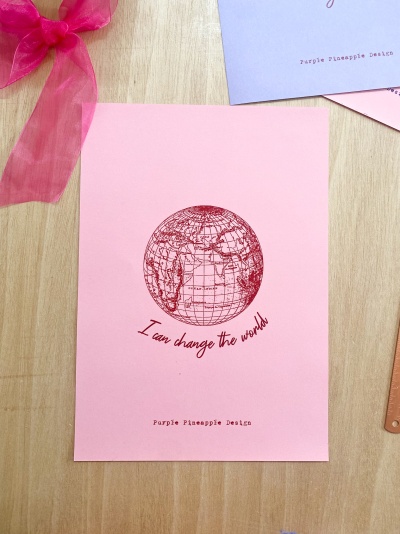 Cartão rosa com desenho de globo e texto motivacional em vermelho colocado sobre mesa de madeira clara