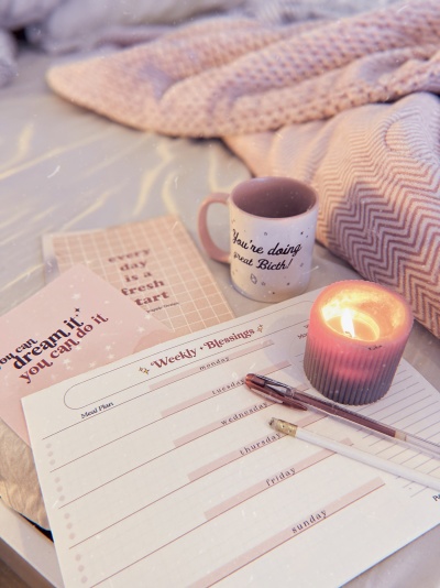 Ambiente com cobertor rosa, caneca com frase, vela acesa, plano semanal e cartões motivacionais.