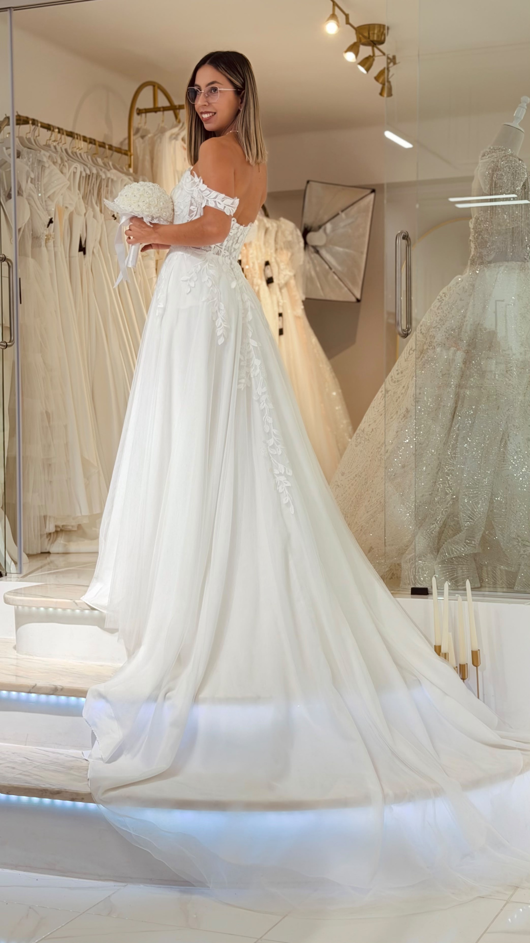 Vestido Amarílis Vestido de noiva branco com renda floral e saia ampla, mulher a segurar bouquet branco em loja