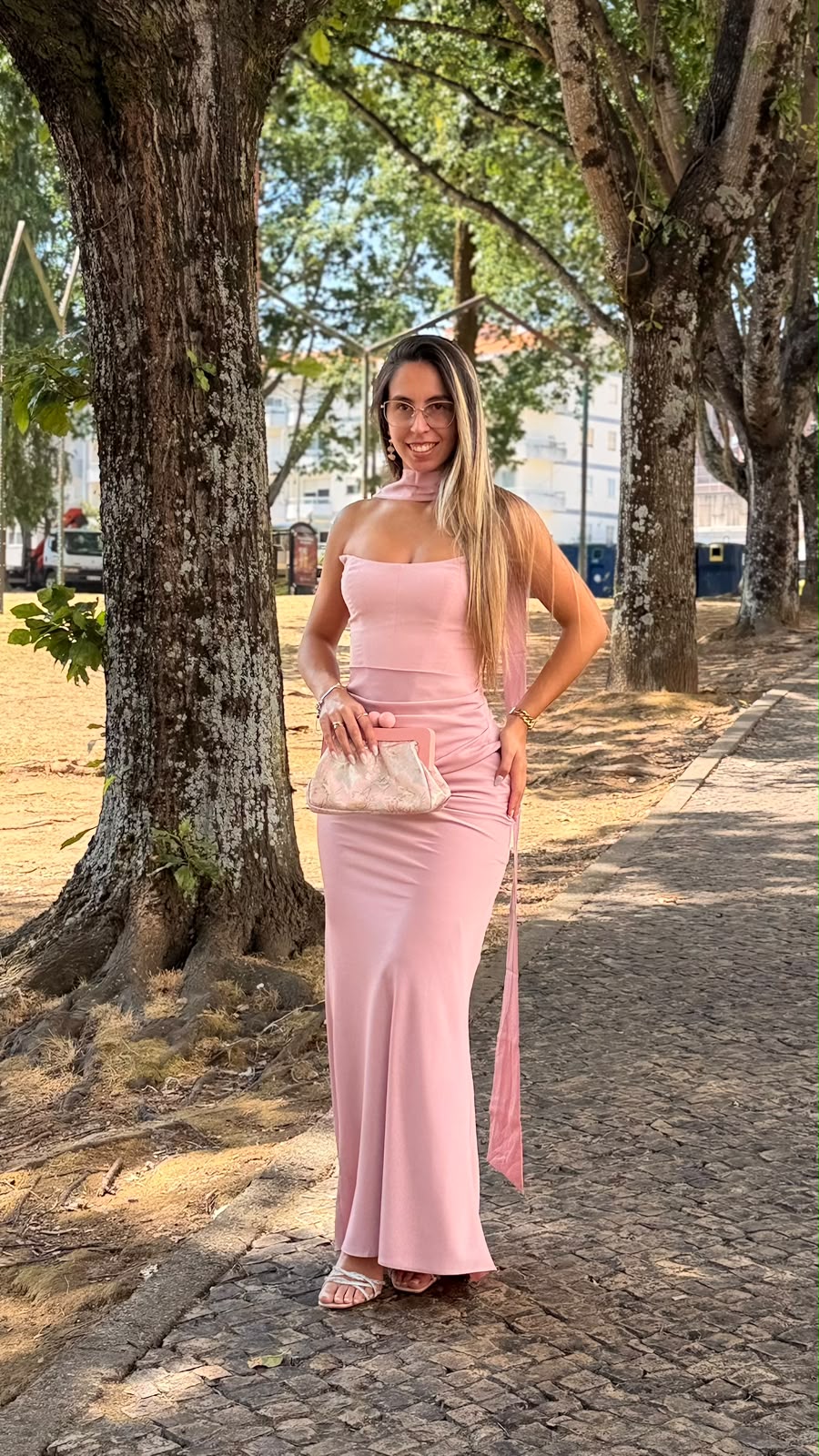 Vestido Romana Mulher com vestido longo rosa e mala bege segurando, em rua com árvores.