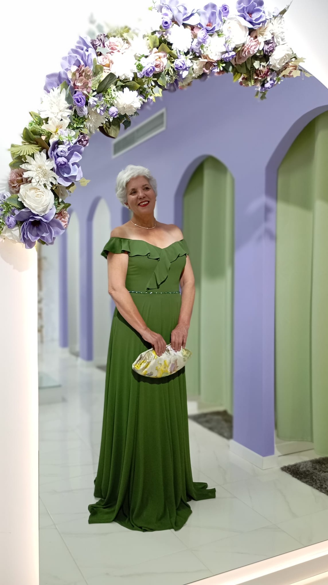 Vestido Nataly Mulher com vestido verde posando em arco floral em ambiente com paredes lilás