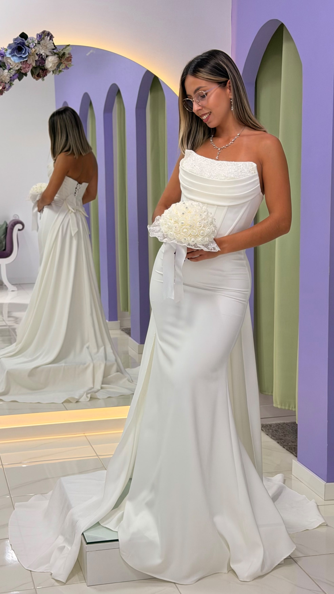 Vestido Éden Noiva Vestido de noiva branco com corpete drapeado e saia longa, mulher segurando bouquet branco em ambiente com espelho