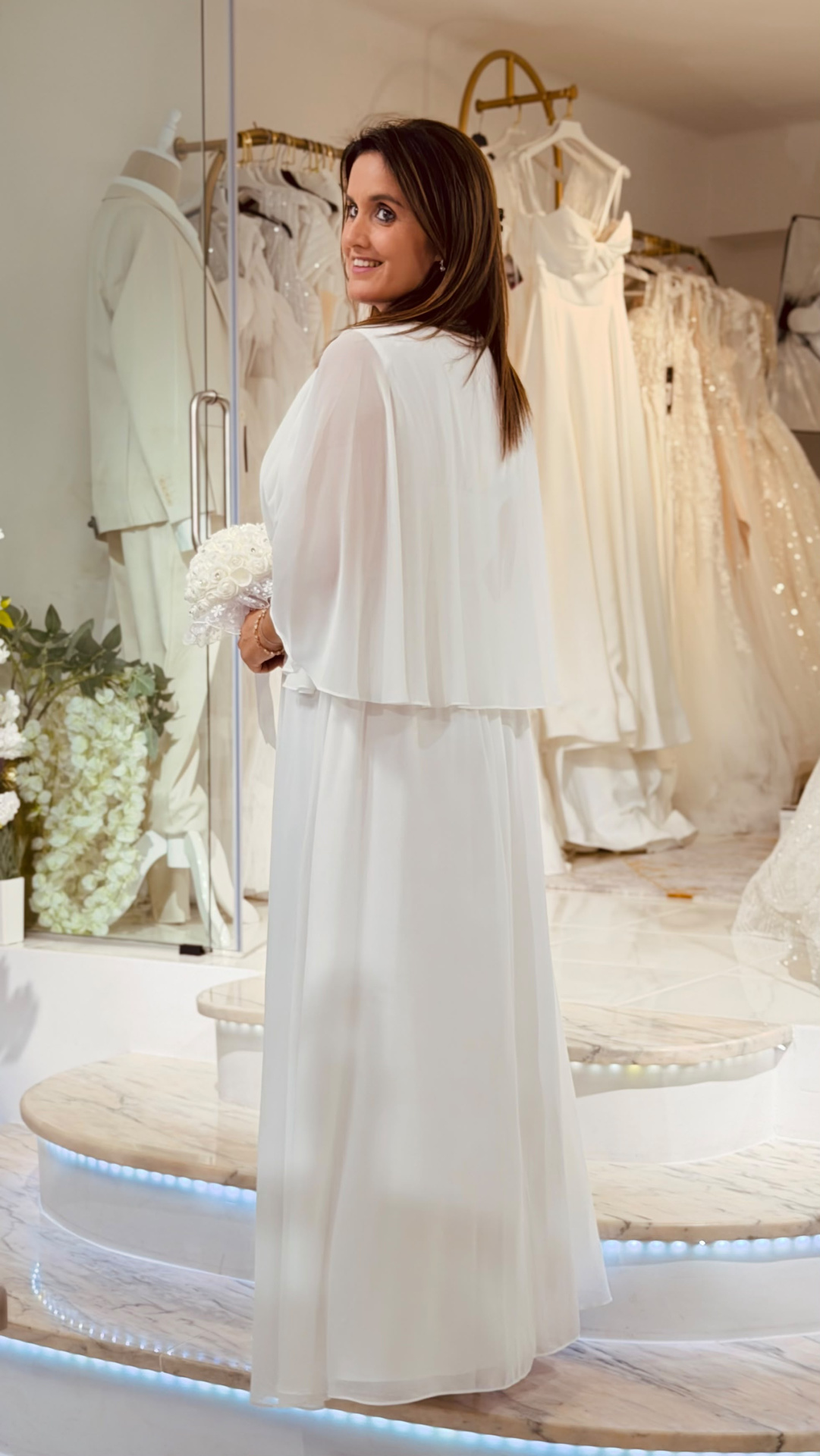 Vestido Rosário Noiva Mulher de costas a usar vestido de noiva branco com capa curta, segurando bouquet, em loja de vestidos de noiva.