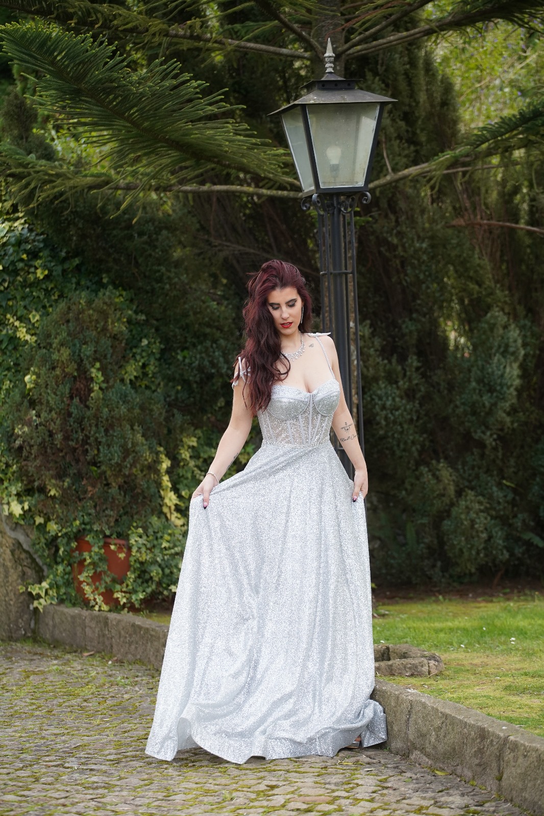 Vestido Princess Glow Mulher com vestido longo prateado brilhante em ambiente exterior com poste de luz e vegetação