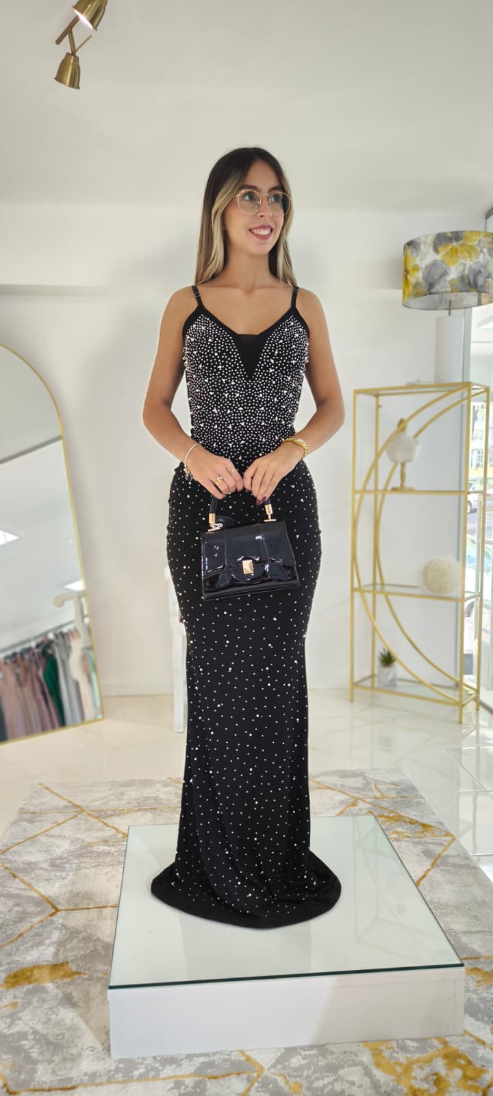 Vestido Tendy c/Pérolas Mulher com vestido longo preto de bolinhas brancas e mala preta de mão num ambiente interior moderno.