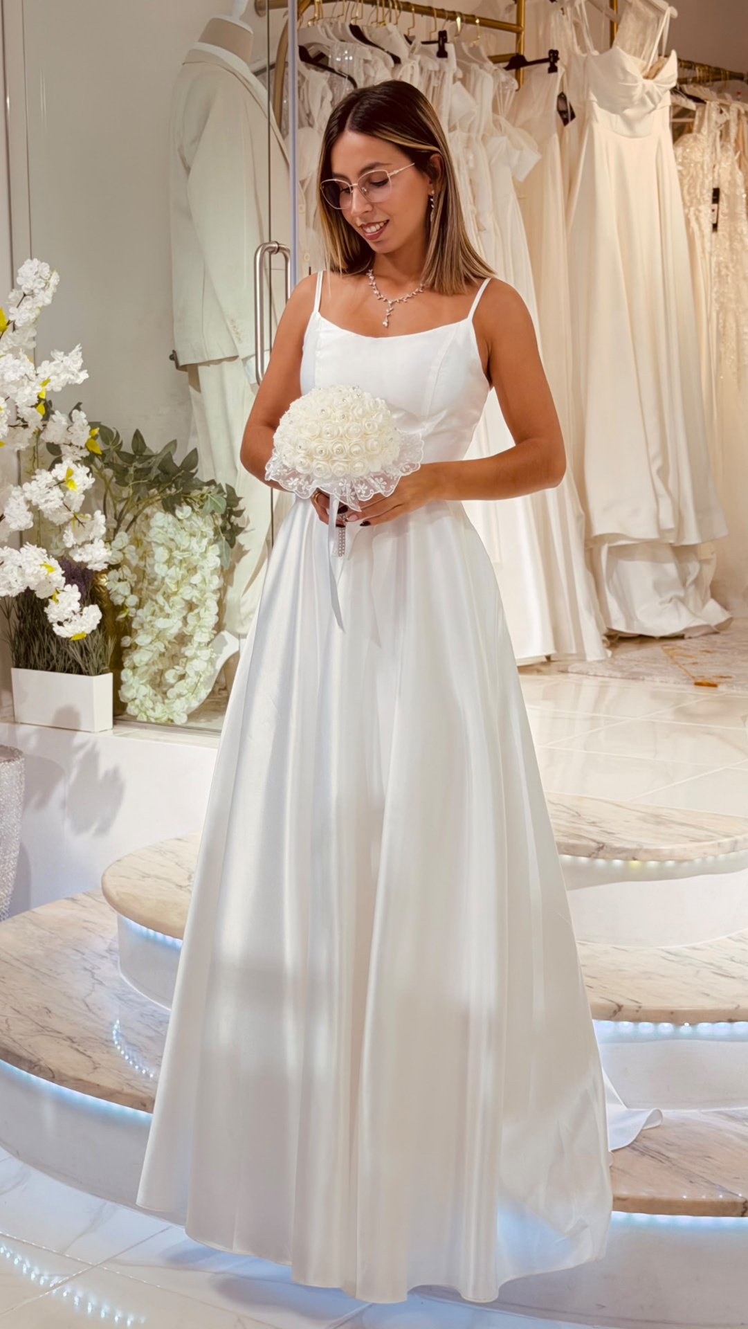 Vestido Helene Noiva Vestido de noiva branco acetinado com alças finas, bouquet branco, mulher na loja de vestidos
