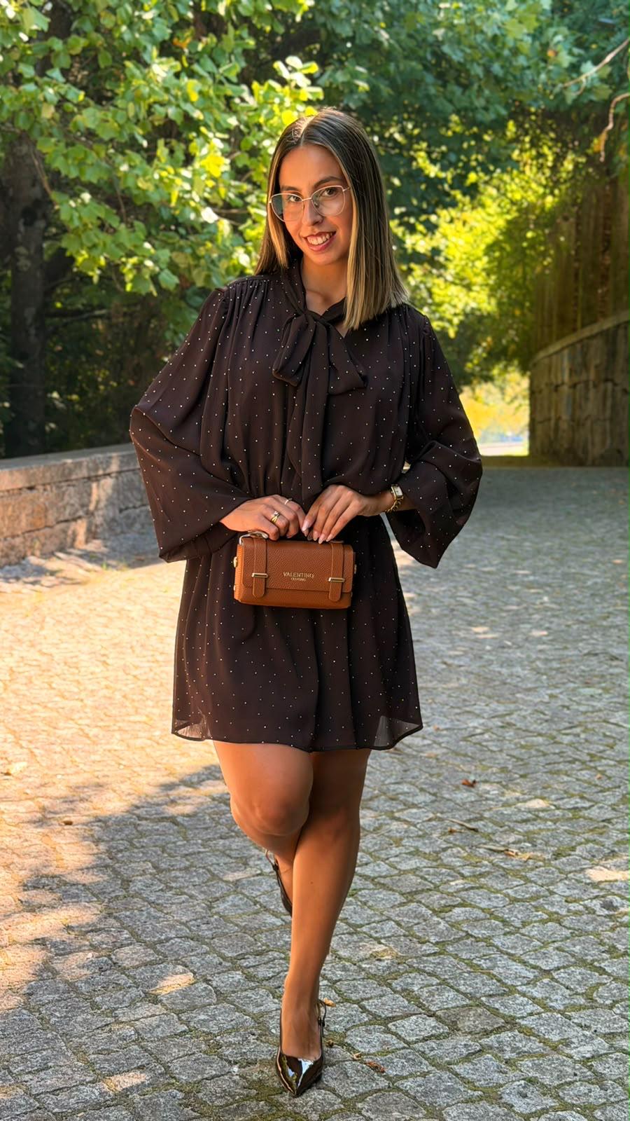 Vestido R&L Castanho Mulher com vestido preto de bolinhas, sapatos pretos e carteira castanha, em passeio exterior com árvores.