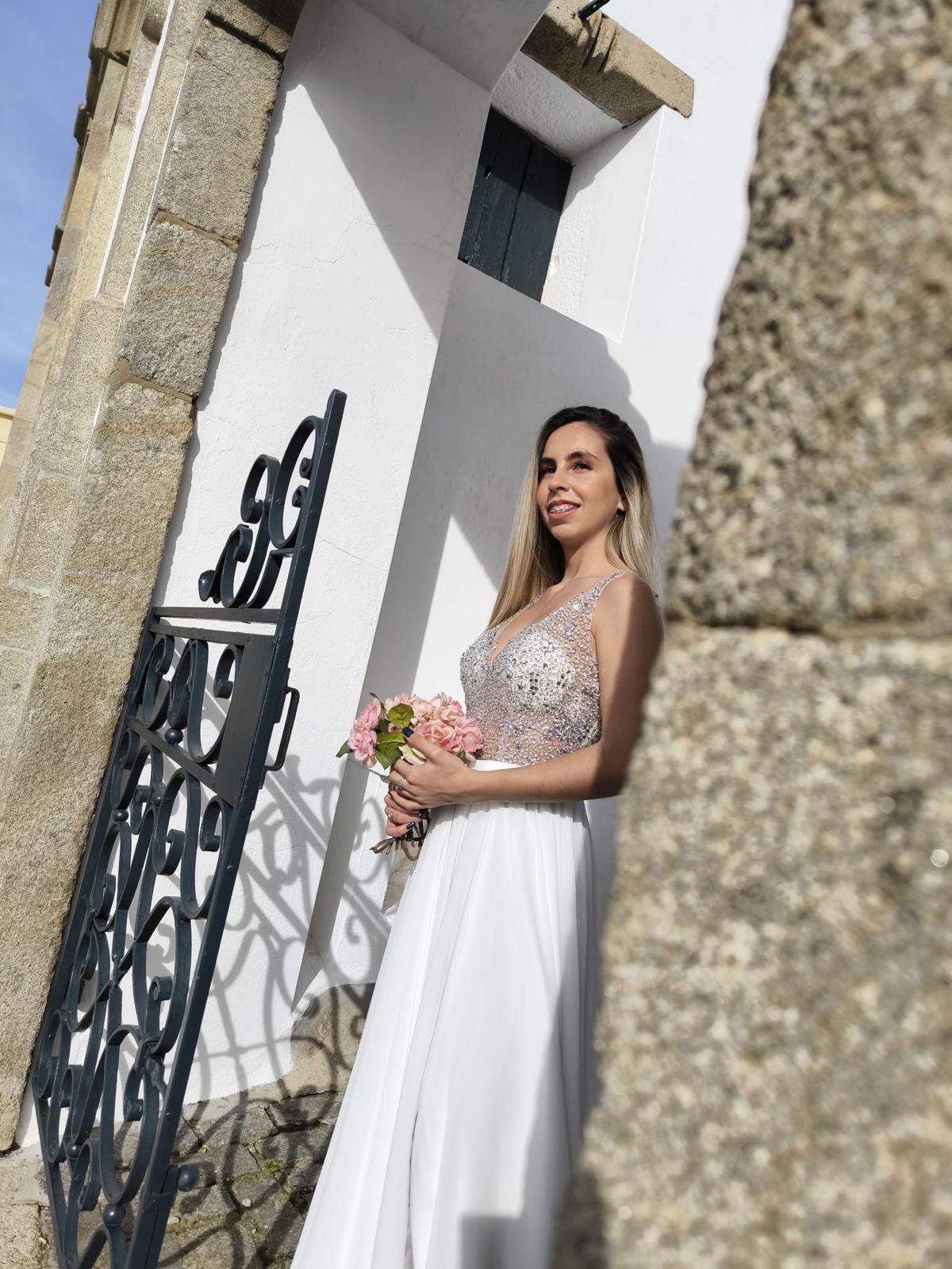 Vestido Simplicidade Serena Mulher com vestido de noiva branco e bouquet de flores junto a portão de ferro preto e parede branca