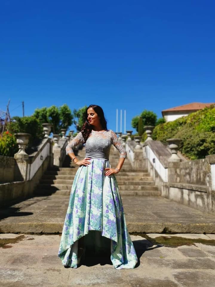 Vestido Mizuky Mulher a usar vestido de cerimónia com parte superior em renda cinza e saia floral assimétrica, em ambiente exterior com escadaria