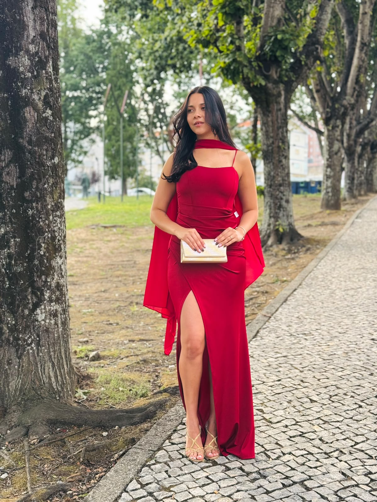 Vestido HUNGARO Mulher com vestido longo vermelho e carteira dourada, em passeio urbano com árvores.