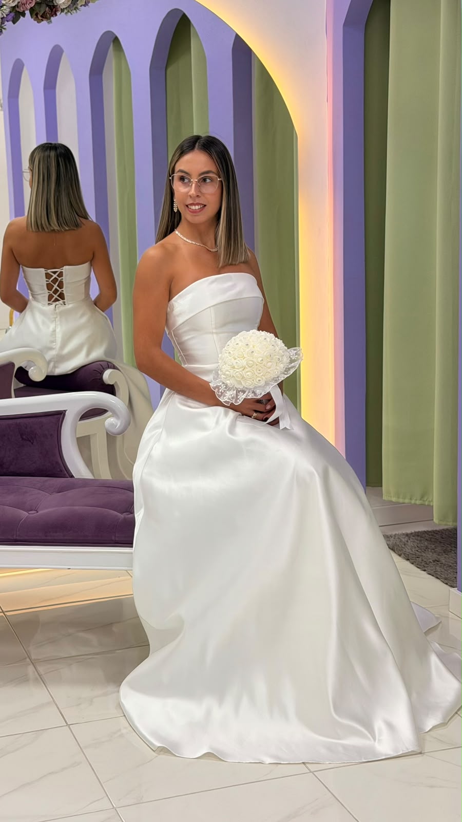 Vestido Alizee Noiva Mulher de vestido de noiva branco acetinado sentada com ramo de flores brancas num ambiente com paredes lilás e verde