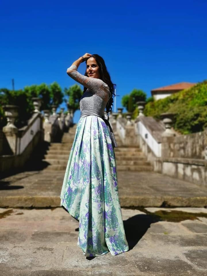 Vestido Mizuky Mulher a usar vestido longo com parte superior prata brilhante e saia florida verde, lilás e branco ao sol em escadaria de pedra.