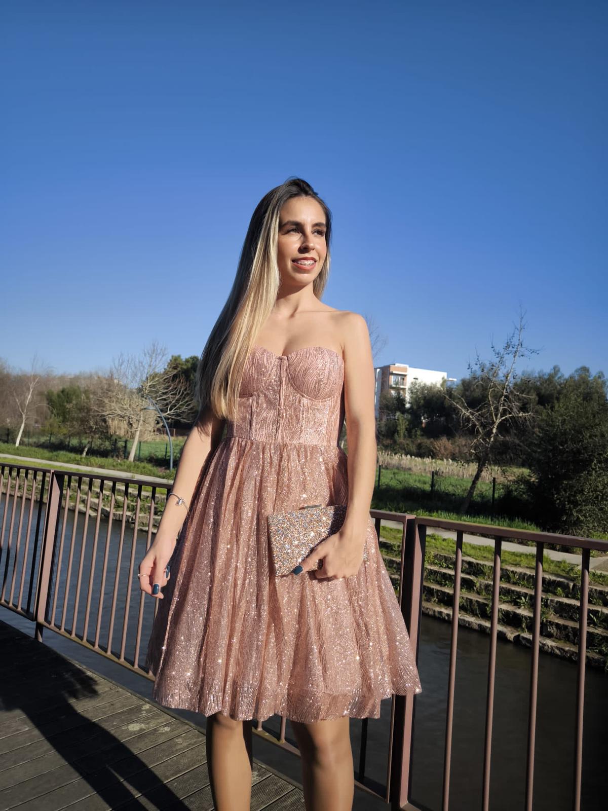 Vestido Glam Princess Mulher com vestido rosa cintilante segurando clutch prateada ao ar livre numa ponte