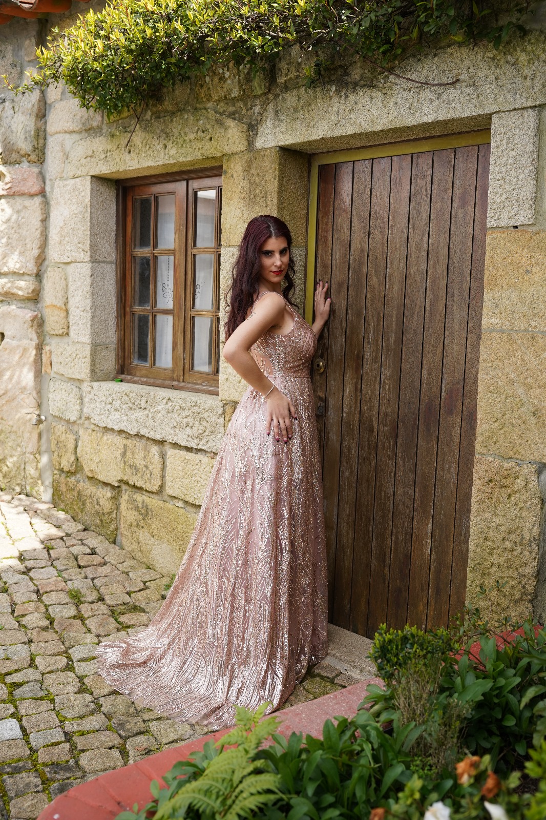 Vestido Camila Glow Vestido de festa longo rosa brilhante junto a porta de madeira e parede de pedra