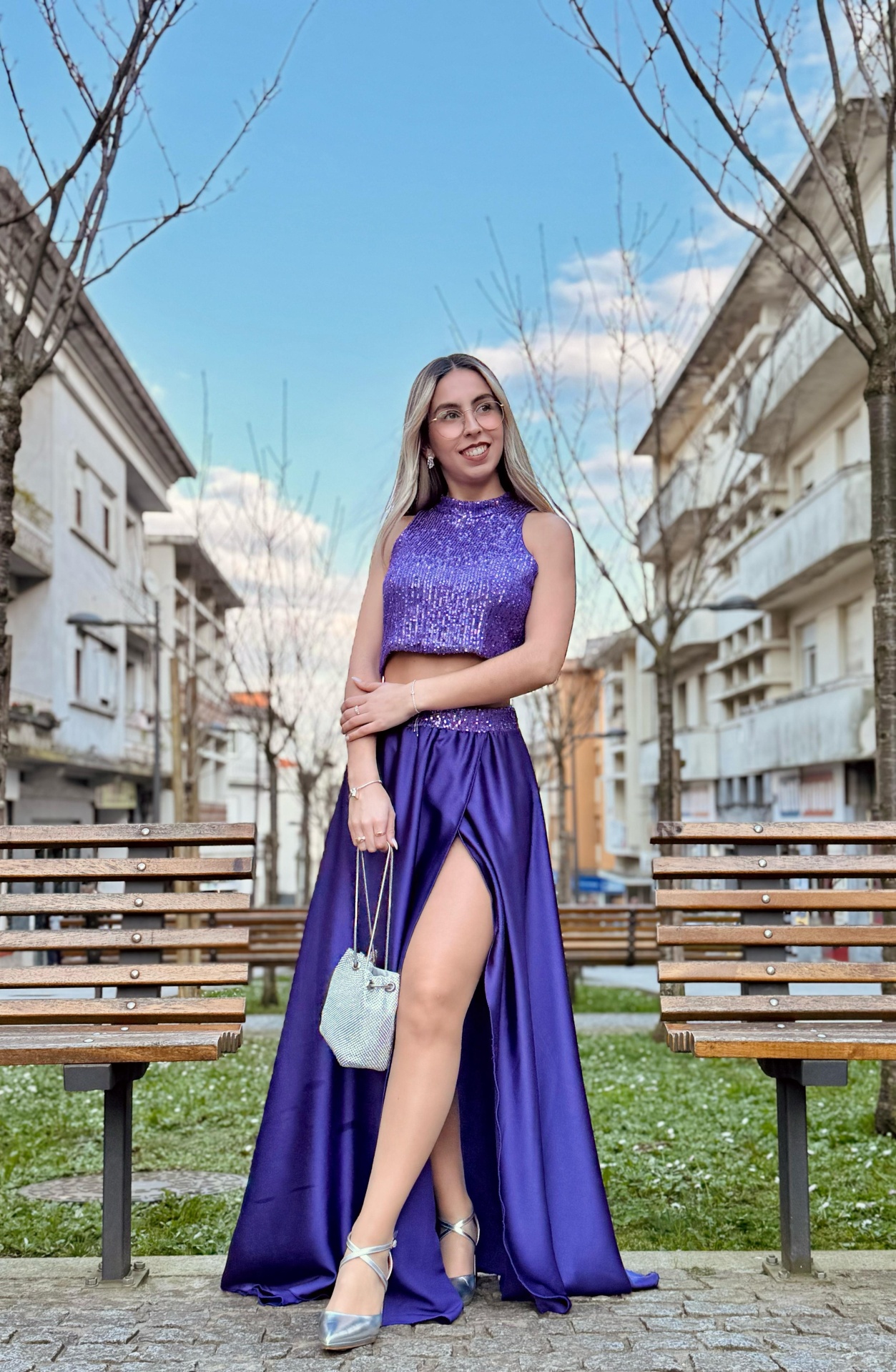 Conjunto Irina Mulher com vestido roxo de duas peças e mala branca em ambiente urbano com bancos de madeira.