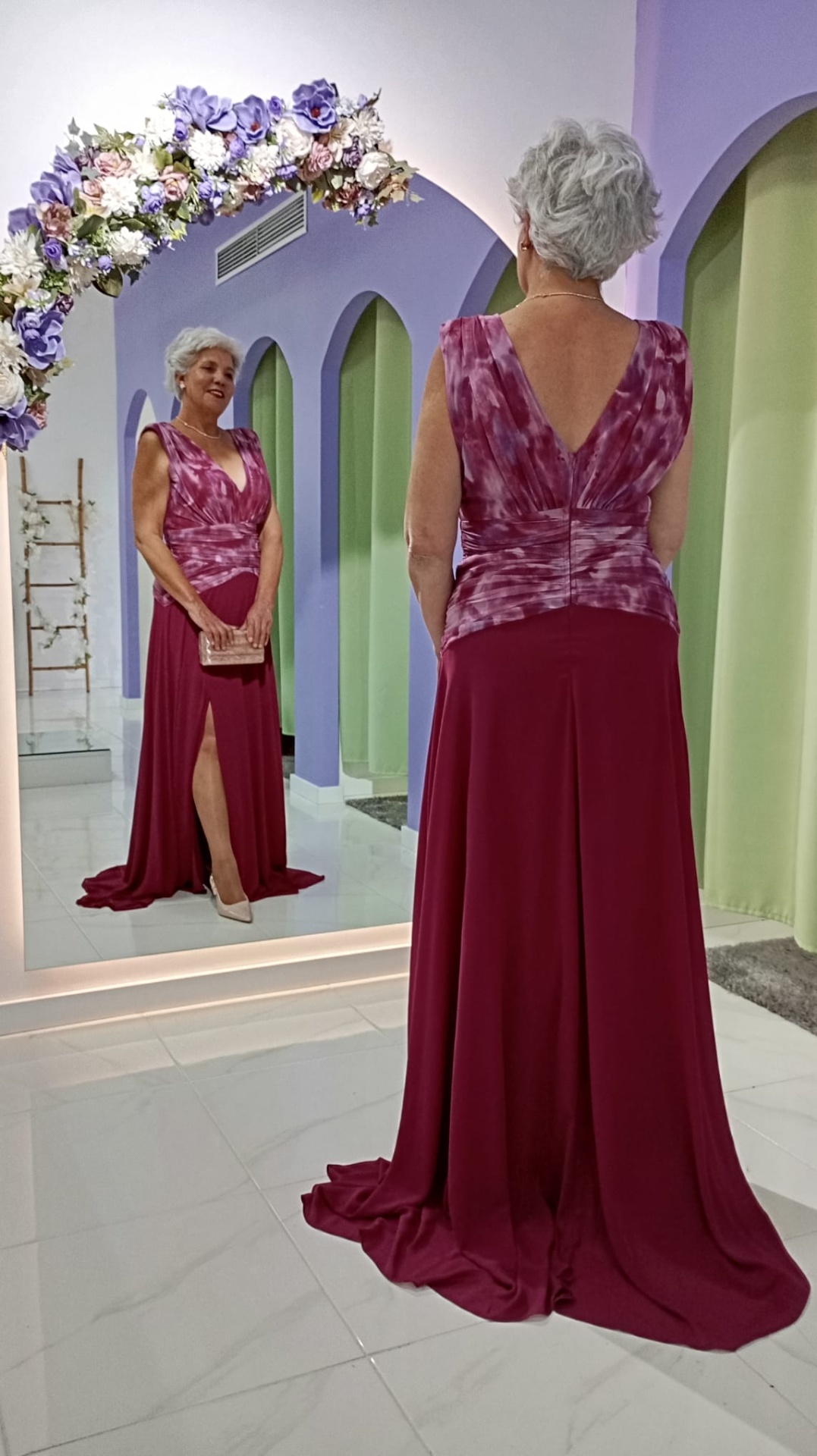 Vestido Andore Plus Size Vestido longo vermelho com parte superior estampada em tons avermelhados e detalhe de racha, mulher em espaço com paredes lilás e arco de flores