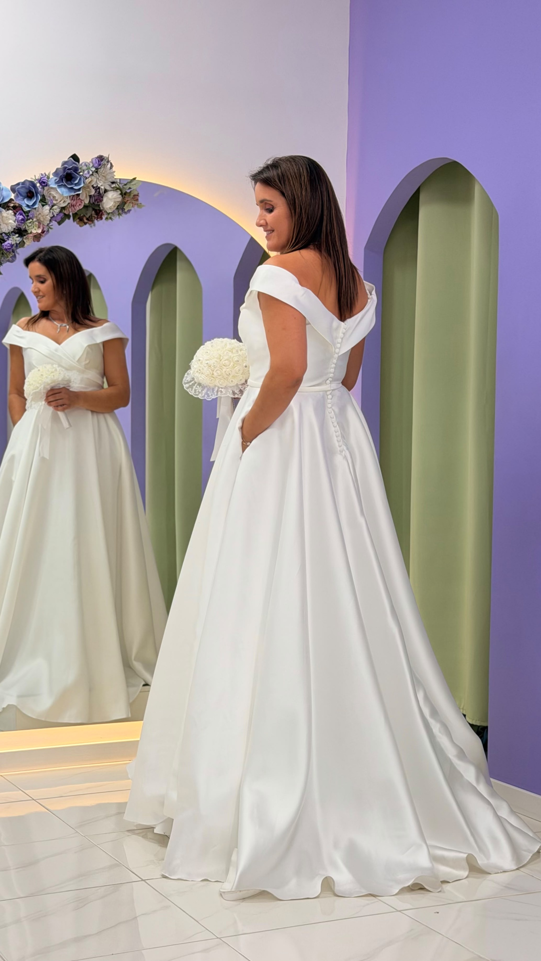 Vestido Gabrielle Noiva Mulher com vestido de noiva branco refletida em espelho com flores azuis, arco roxo ao fundo