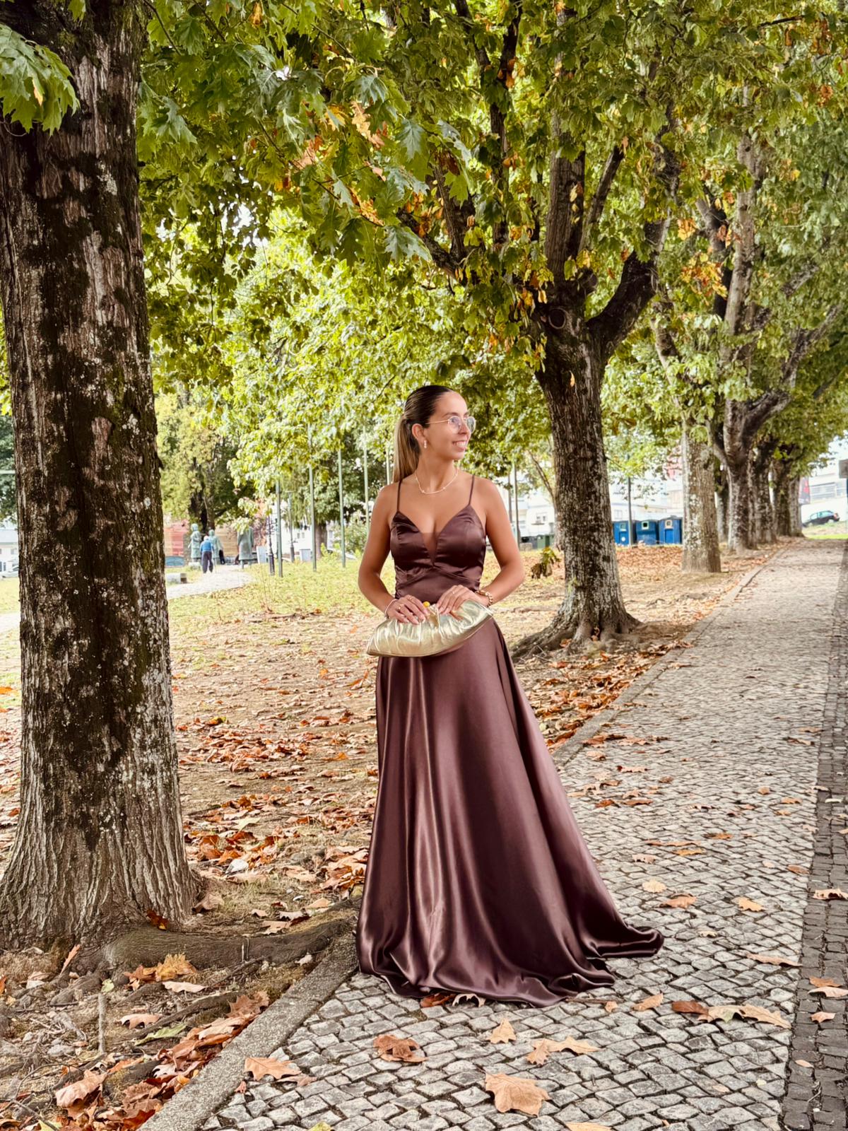 Vestido Doriane Mulher com vestido longo de cetim castanho e carteira bege em passeio com árvores