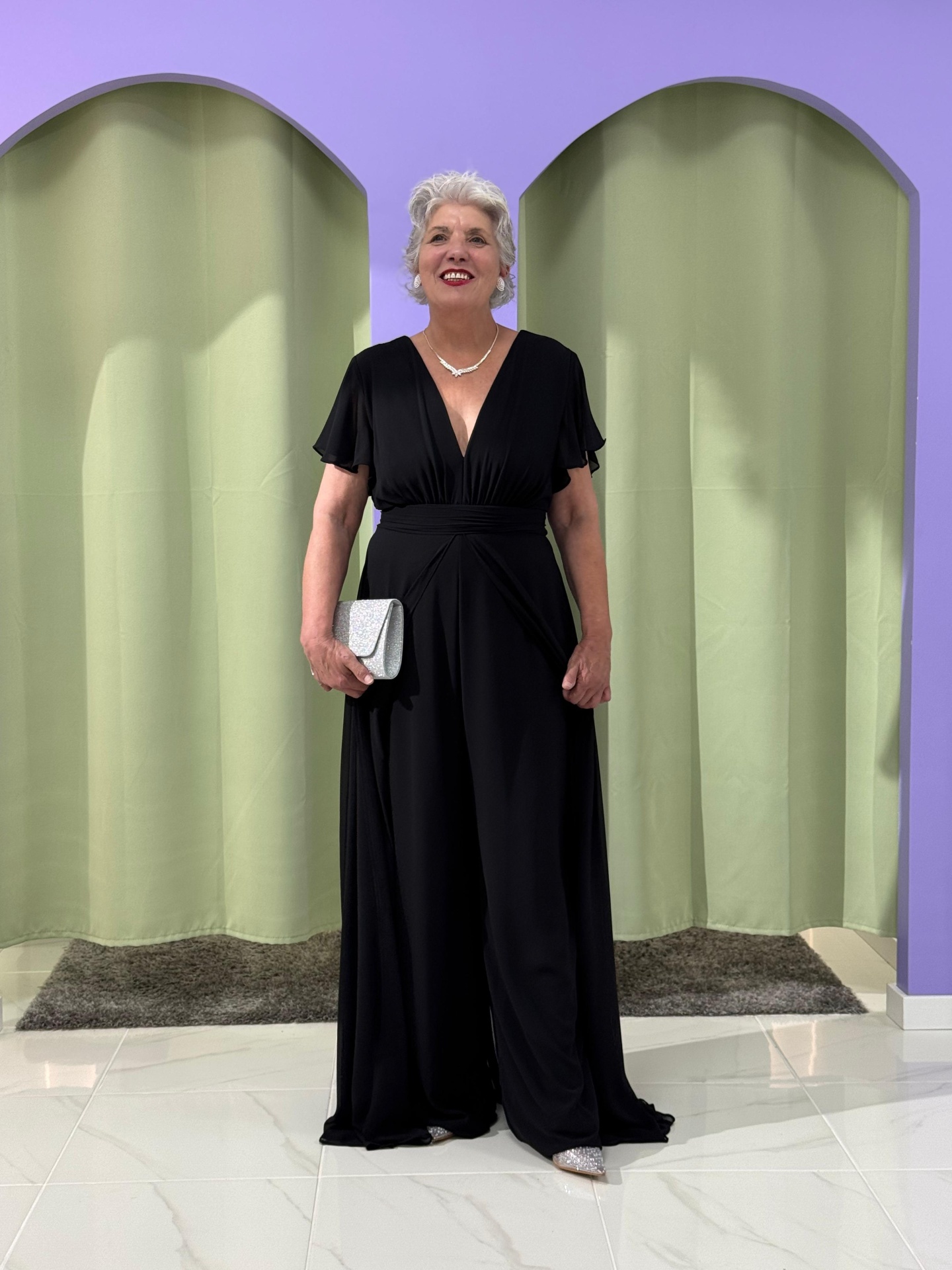 Macacão/Vestido Plus Size Mulher sénior vestindo macacão preto e segurando clutch cinza prata em ambiente com cortinas verdes e parede lilás.