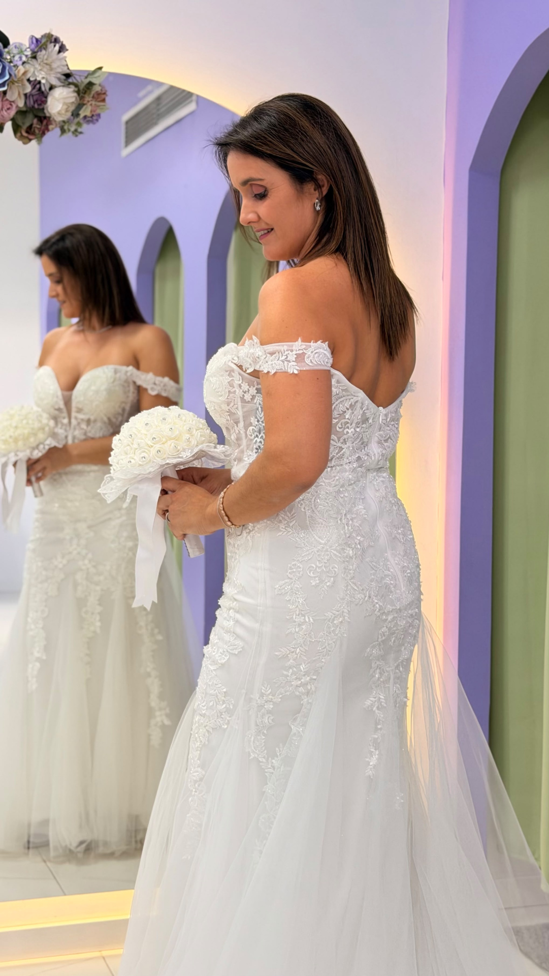 Vestido Wedding Ilusion Mulher com vestido de noiva branco rendado segurando ramo branco em frente a espelho